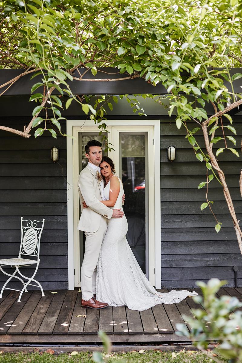 KWH real bride Emma and will stand in front of their black wedding bungalow. She wears the Rylie gown, a lace fit and flare wedding dress with traumpet train.