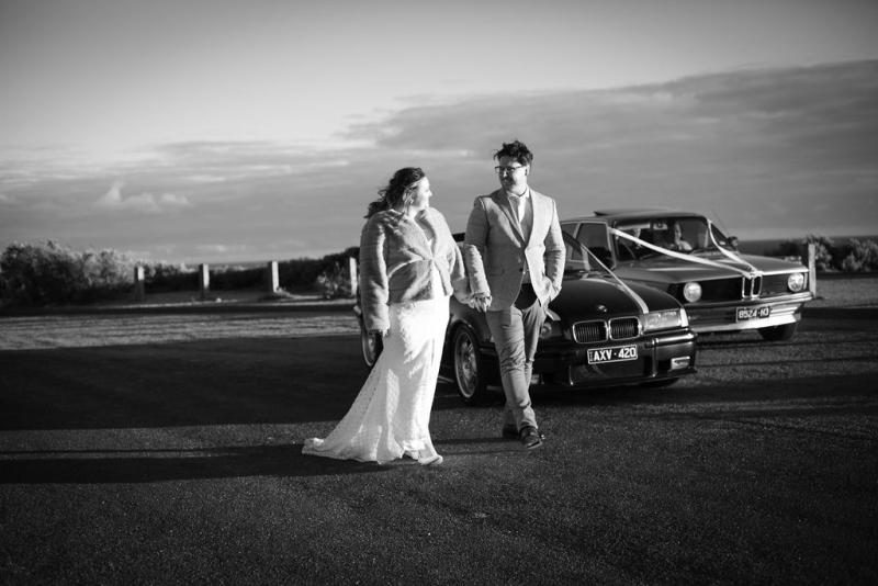 KWH real bride Meg and Tom stand in front of BMW's. She wears her Bobby gown paired with a fur cape.