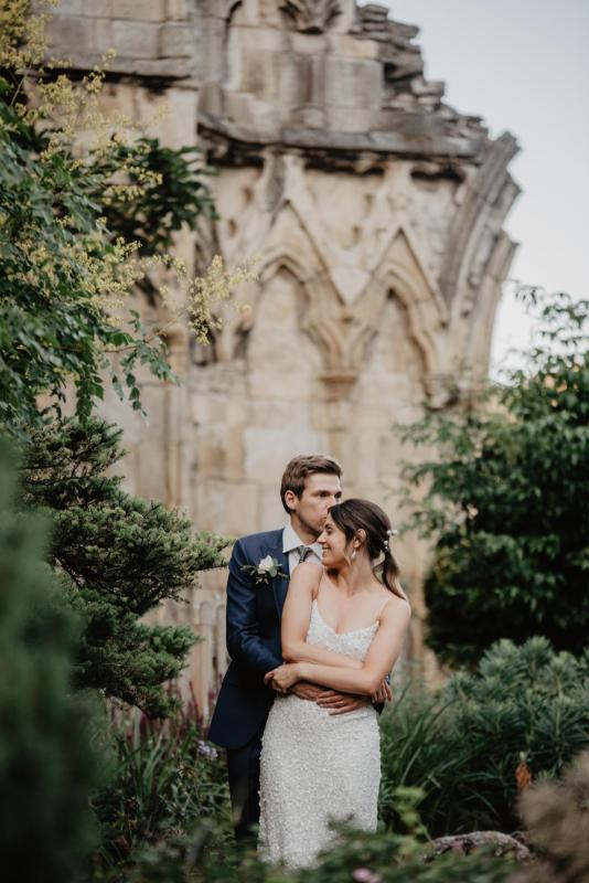 KWH real bride Monica and James hug in the castle gardens. She wears the classic Anya gown, a modern sequin wedding dress with thin straps.