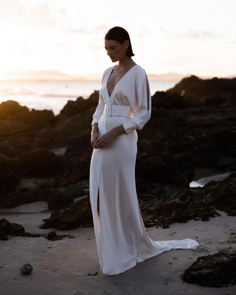 The Margo gown by Karen Willis Holmes, a V-Neck, long sleeve silk satin wedding dress with a fit and flare shape and a split skirt. Model stands against black ocean stones