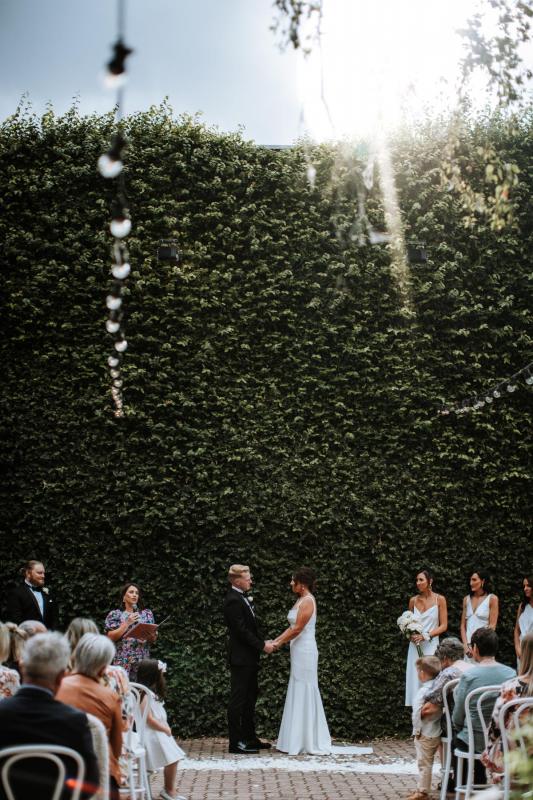 KWH real bride Emily and Dan stand at the alter exchanging vows in front of a large hedge wall at Butler Lane. Emily wears the effortless Violet gown.