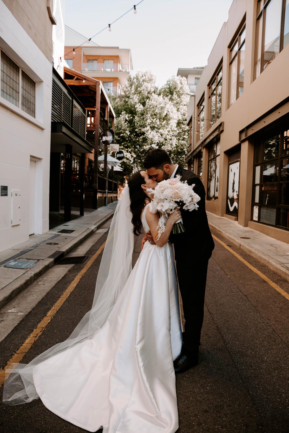 KWH real bride Shenea kissing in the streets with her new husband Lain. She wears the simple elegant Taryn Camille gown by KWH.
