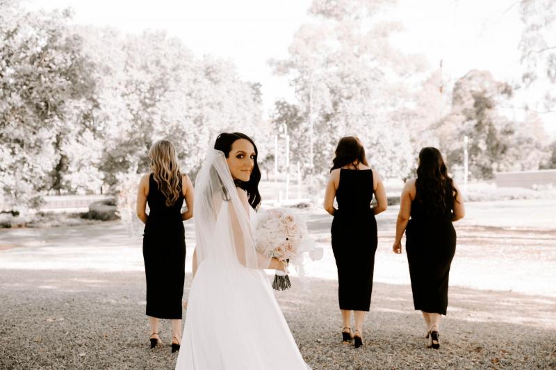 KWH real bride Shenea standing in front of her bridesmaids who wear black. She wears the ivory timeless Taryn Camille gown.