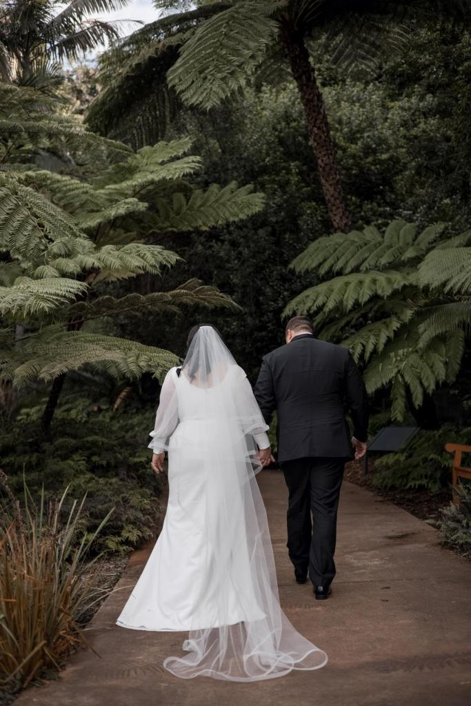 KWH real bride Dani and Thomas walking thorugh a fern forest, she wears the classic Nikki wedding dress with long sleeves and v-neck.