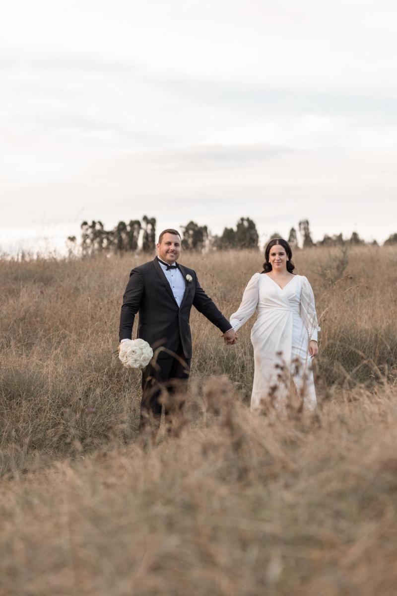 KWH real bride Dani holds hands with Thomas in the field. She wears the classic and simple Nikki wedding dress
