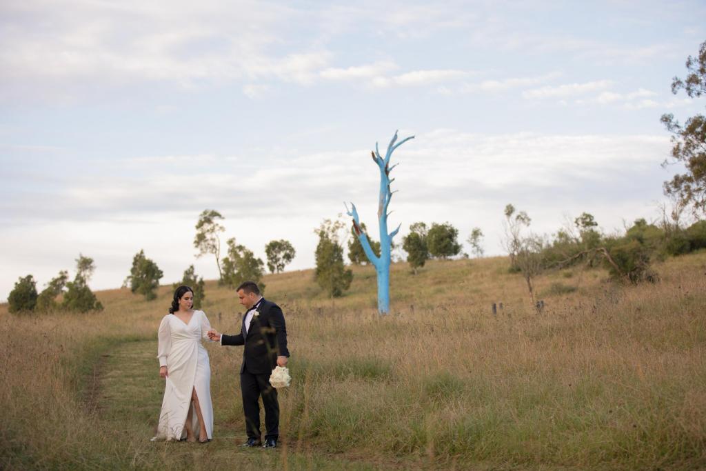 KWh real bride Dani stands in a field by a painted blue tree. She wears the modern curve Nikki wedding dress