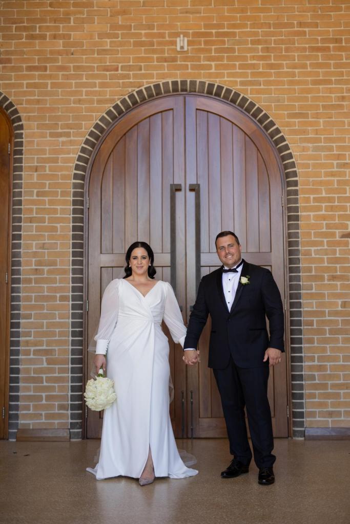 KWH real bride Dani and Thomas stand in front of a arched doorway in her classic Nikki wedding dress.