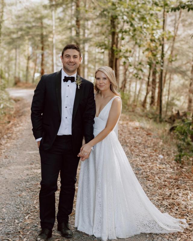 Real bride Megan walking with husband, wearing the Nadia gown; a lace wedding dress for the bohemian bride, featuring a V-neck and side split.