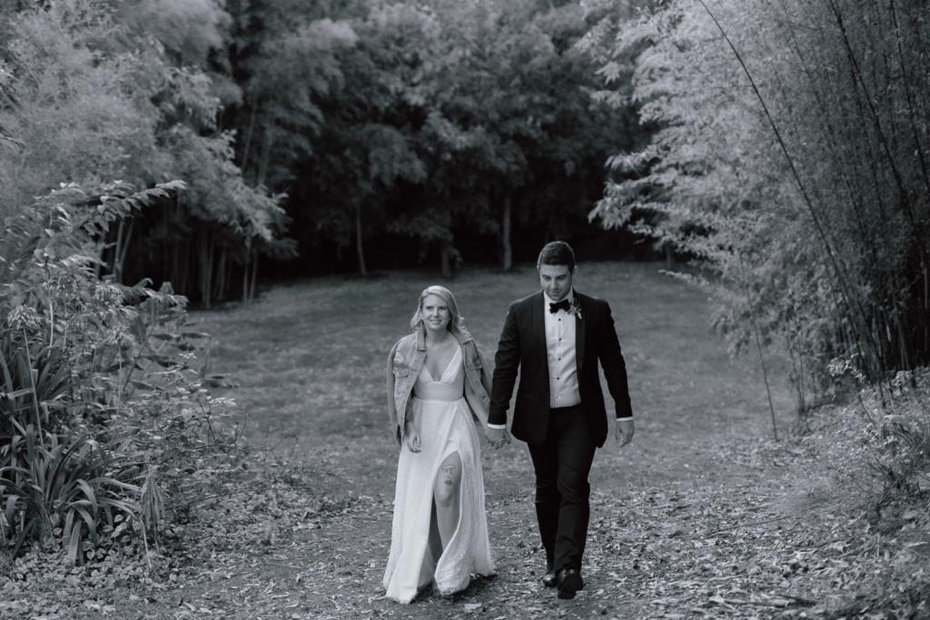 Real bride Megan walking with husband, wearing the Nadia gown; a lace wedding dress for the bohemian bride, featuring a V-neck and side split.