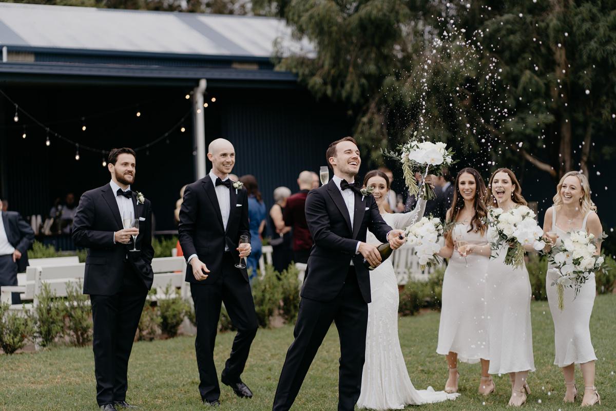 Karen Willis Holmes bride Anna's husband Mitch popping the champagne surrounded by the wedding party; wearing the Cassie wedding dress.