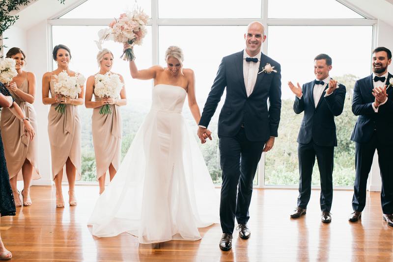 KWH bride Samantha and husband Adam walking down the aisle. Couple is surrounded by their wedding party as bride celebrates with bouquet in the air. Samantha wears the Esther wedding dress with a fitted skirt and long train.