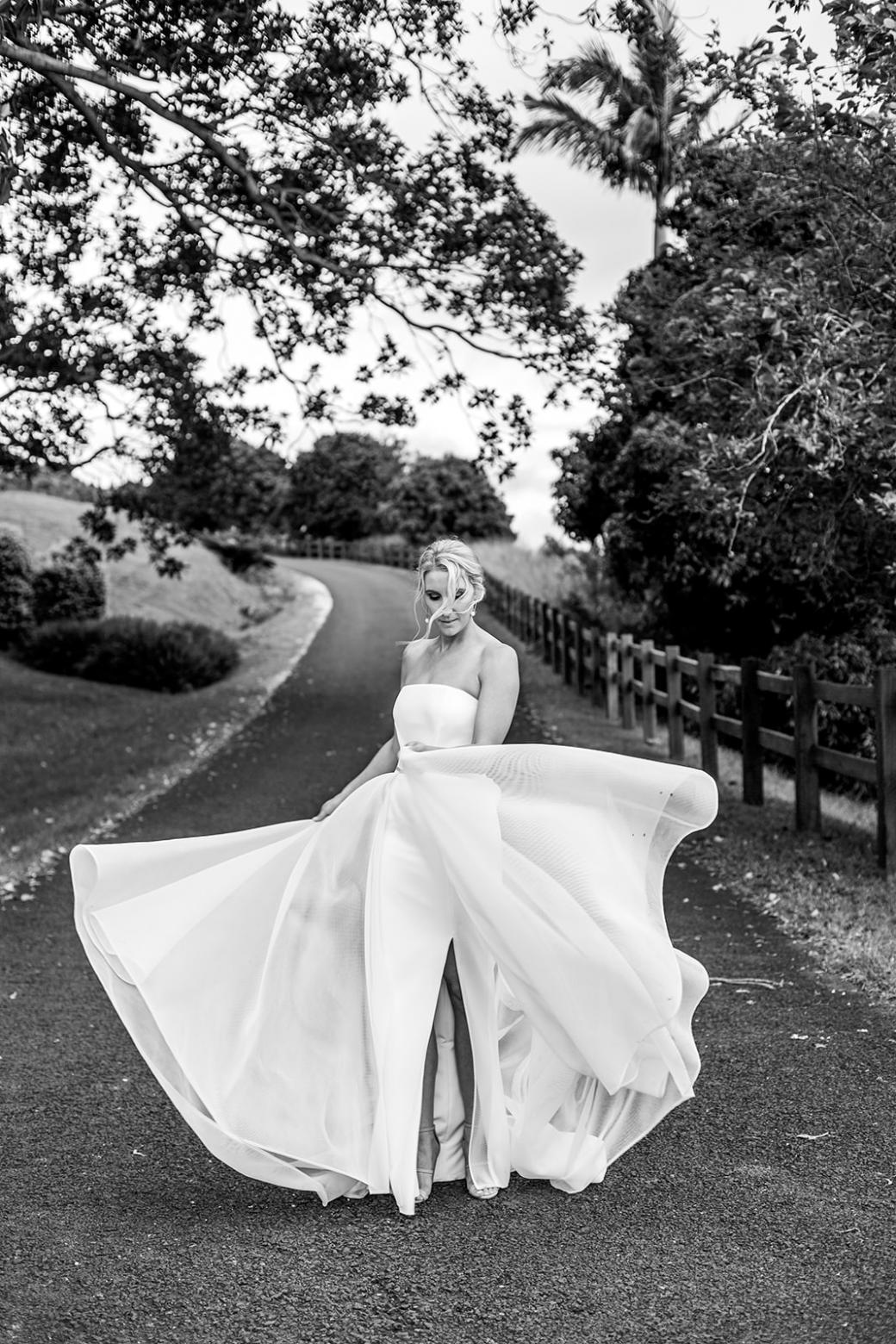 B&W image of KWH real bride Samantha twirling in her Esther wedding dress. The strapless a-line dress features a front split, fitted pencil skirt and long train made out of modern mesh.