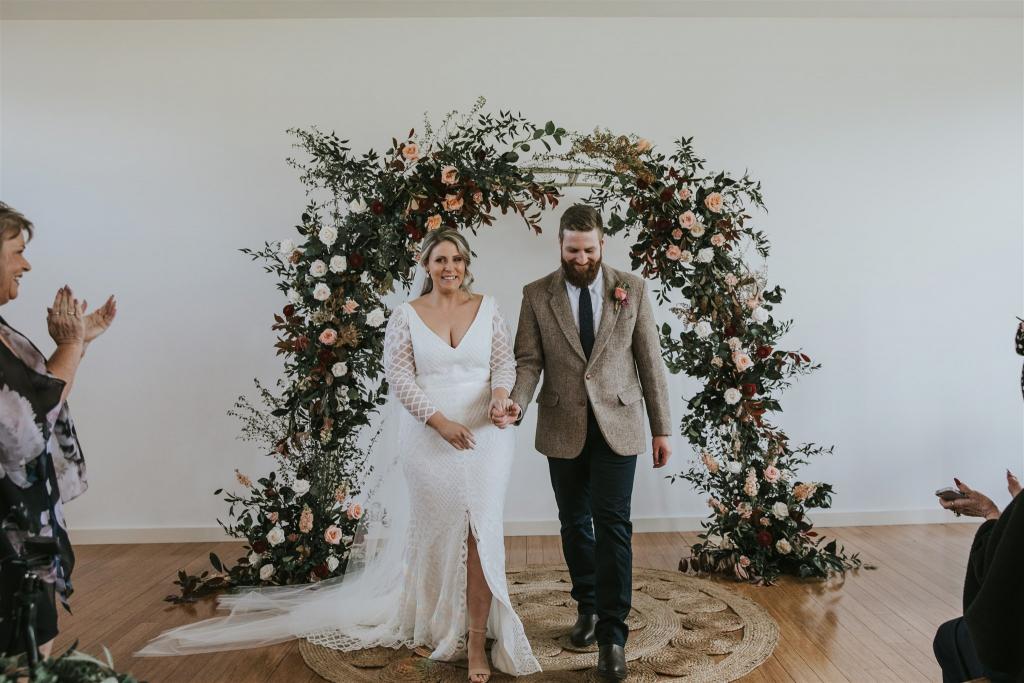 Curvy bride Lucy standing under flower arch at wedding ceremony, bride wearing the Bobby gown; a modern lace wedding dress with long sleeves by Karen Willis Holmes.