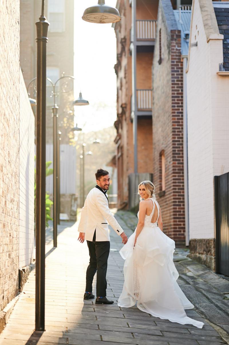 Real bride Claire wears the Cindy gown, a beaded halterneck wedding dress by Karen Willis Holmes to her Sydney wedding, along with the dramatic Diamond detachable trains. The new couple looks back at the photographer as they walk down the street.