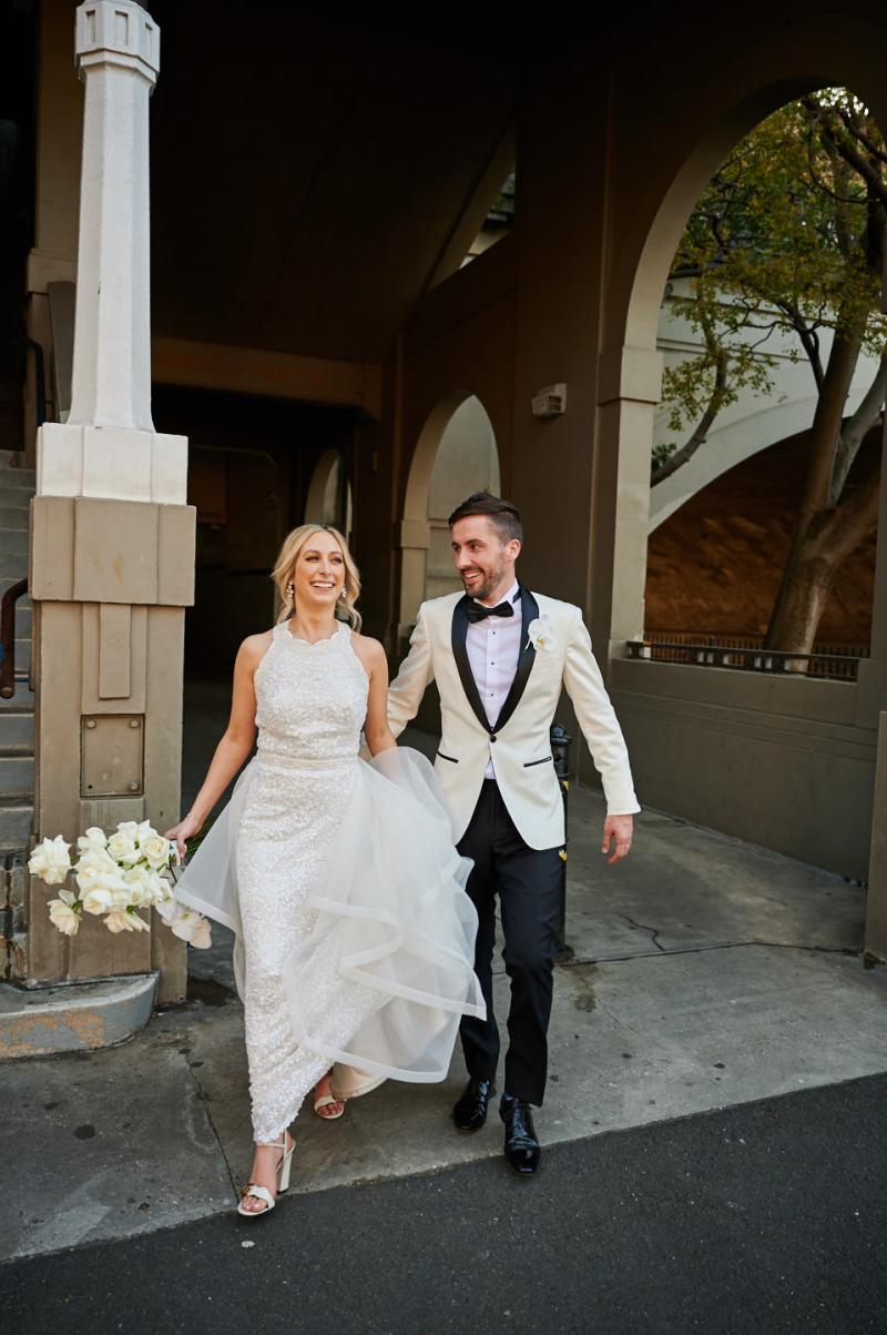 Real bride Claire wears the Cindy gown, a beaded halterneck wedding dress by Karen Willis Holmes. New husband holds her train while walking out of the venue.