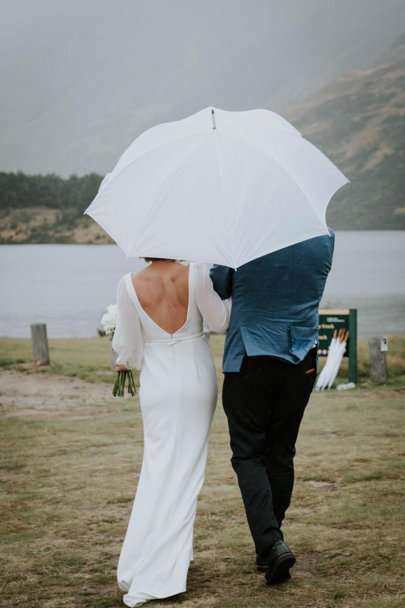 KWH bride Lauren and husband Will under an umbrella. Lauren holds a timeless bouquet and wears the Nikki wedding dress.