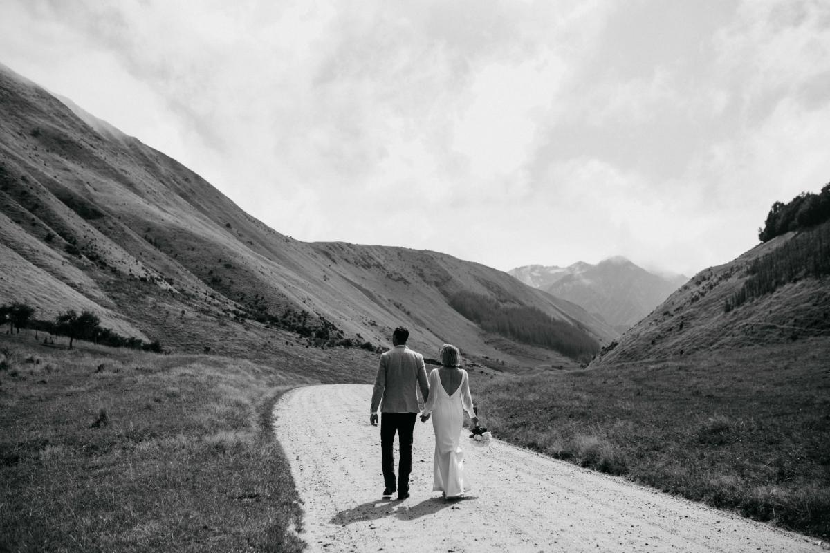 KWH real bride Lauren and Will walk together a in front of magical New Zealand landscape. The bride wears the modern sleeved Nikki gown