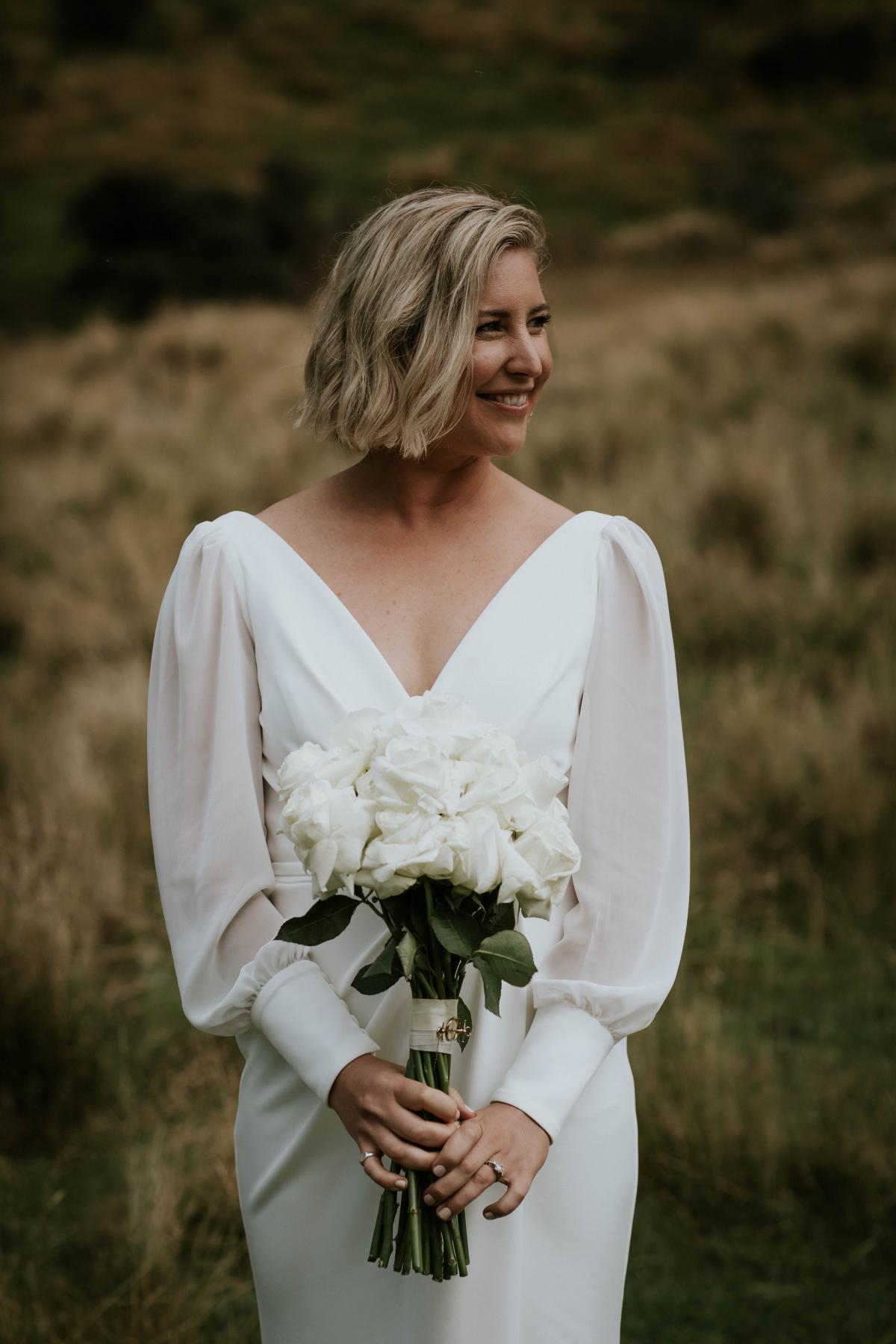 KWH real bride Lauren holding bridal bouquet in Nikki wedding dress. The gown features a wrap style with chiffon sleeves and front split.