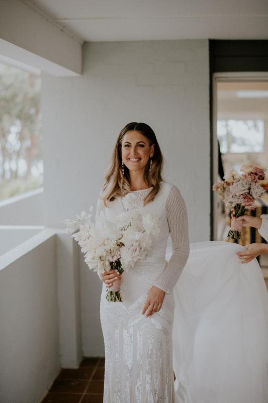 Real bride Penelope wears the Cassie gown while holding a white modern bridal bouquet; a long sleeve art deco beaded wedding dress from the Luxe collection by Karen Willis Holmes.