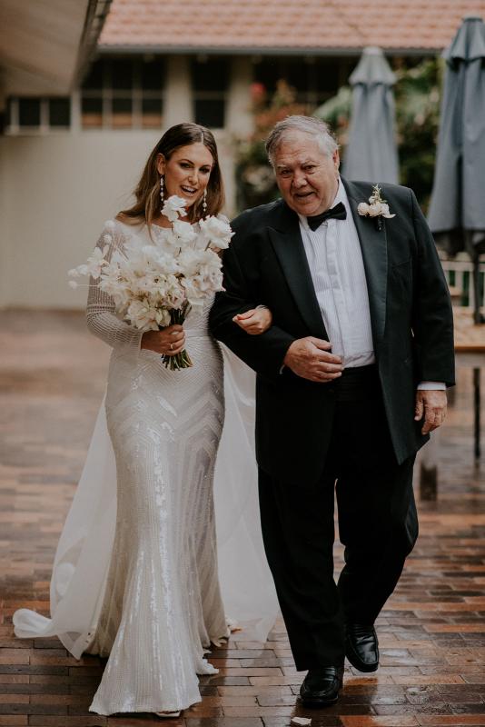 Real bride Penelope is walked down the ceremony aisle by her father, wearing the Cassie gown; a long sleeve beaded wedding dress by Karen Willis Holmes.