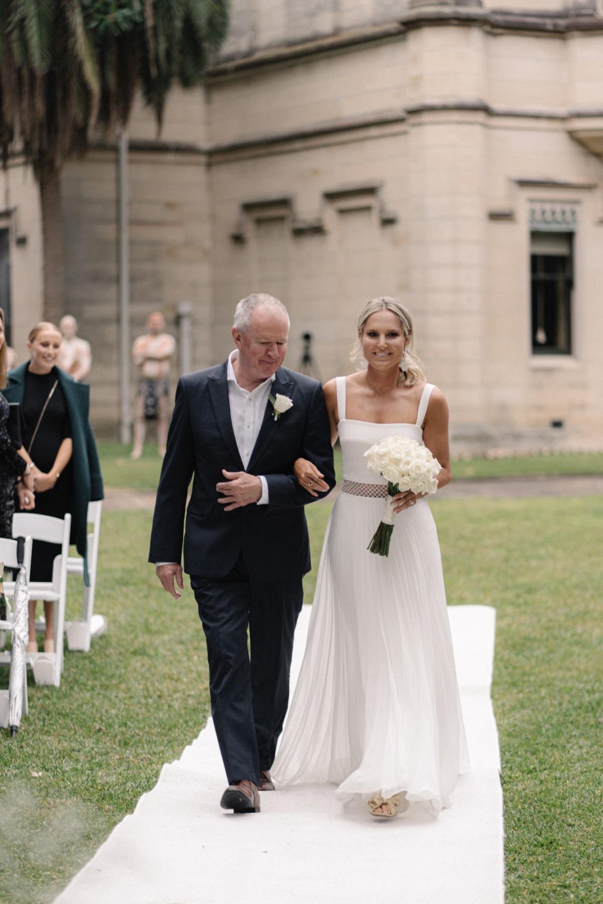 Real bride Maddi walking down the aisle to her garden wedding Indigenous Ceremony, wearing the Daisy gown; a modern wedding dress by Karen Willis Holmes.