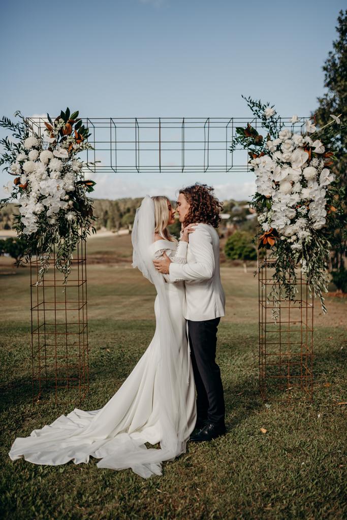 Real bride Hannah kisses husband Rich at wedding ceremony under floral arbour, wearing the Lauren gown with customised long sleeves from the Nikki gown by Karen Willis Holmes