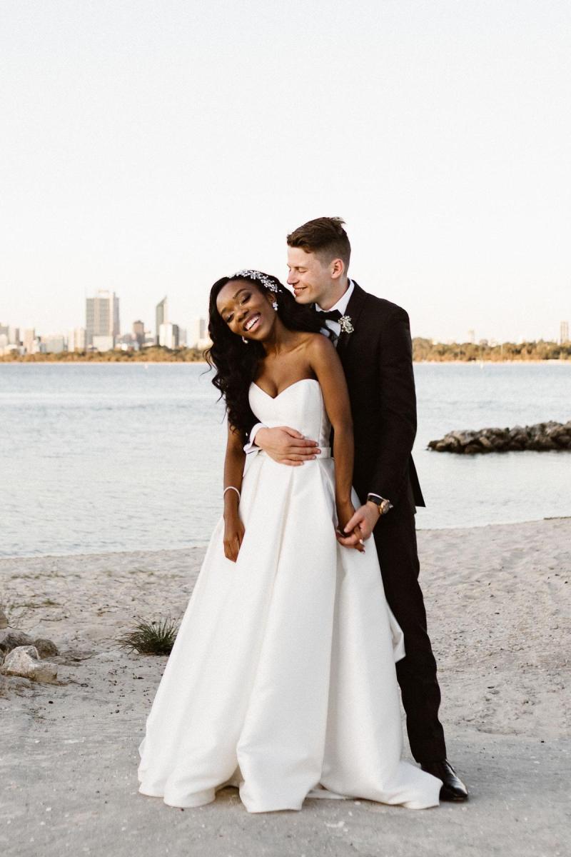 KWH Real bride Ancille and husband Dar stand on the beacha as he wraps his arms around her waist. She wears the timeless Kitty Melanie gown with pockets.