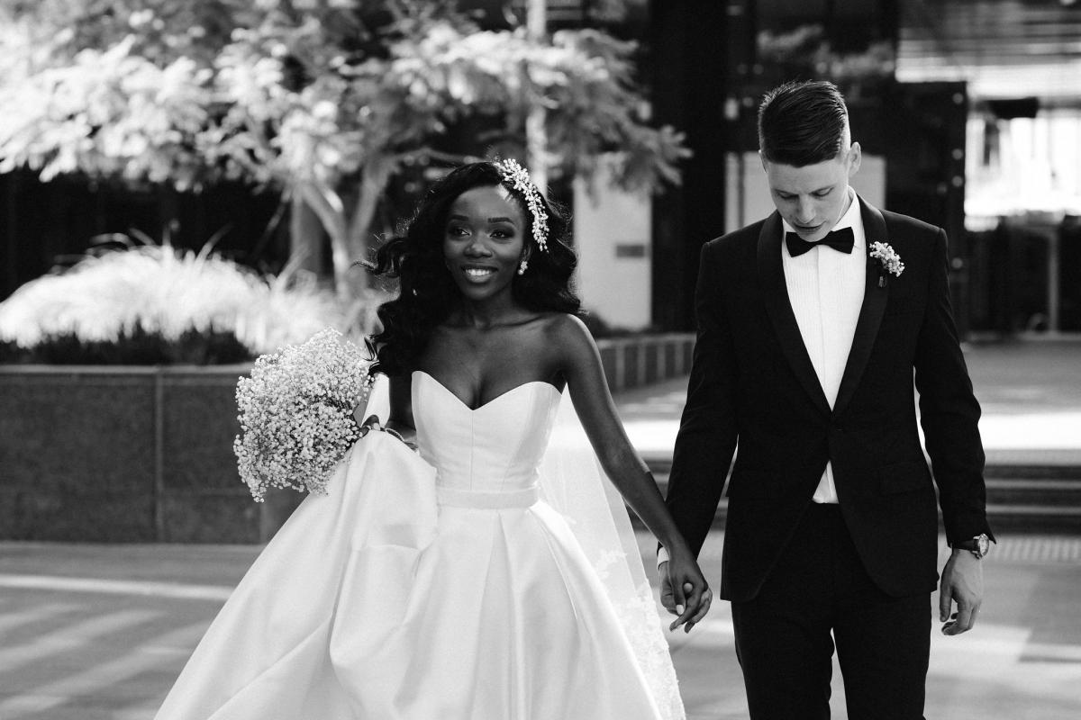 Real bride Ancille and Dar hold hands as they cross the street in this B&W photo. She wears the design your own Kitty Melanie wedding dress from KWH.