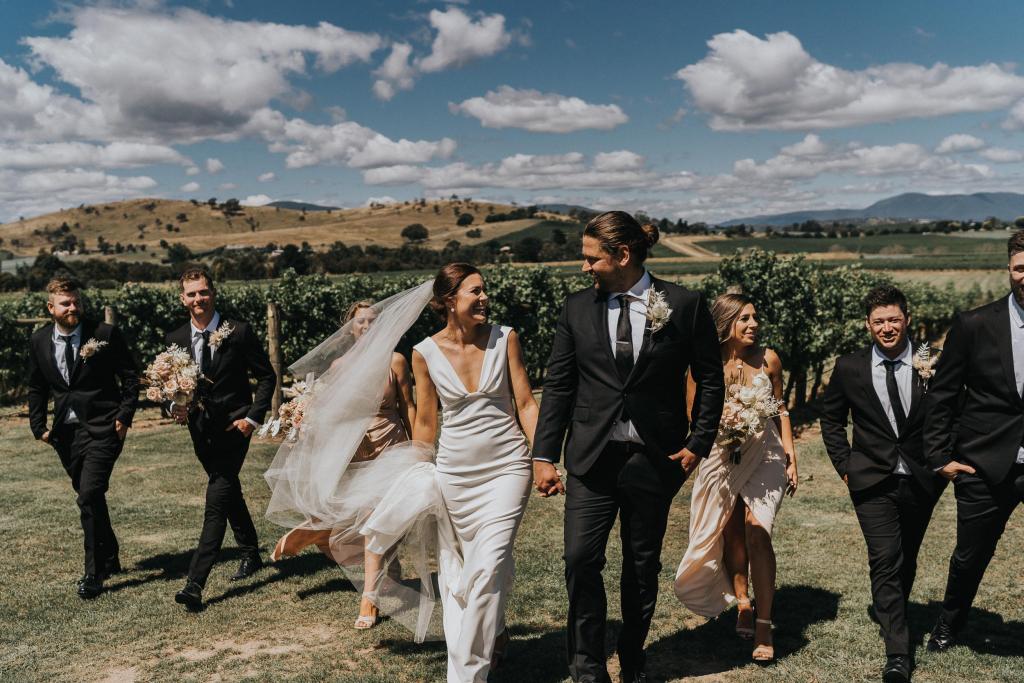 Newly weds, Kate and Mik, walk through the rustic landscape holding hands leading their group of bridesmaids and groomsmen to the reception. Kate wears the plunging v-neck Arabella gown by KWH.