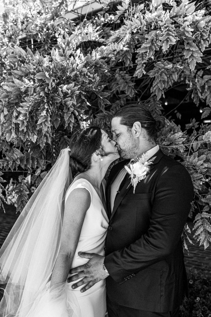 Real bride Kate and Mik kiss in front of a lush bush; she wears the crepe Arbella gown with plunging neckline by KWH.