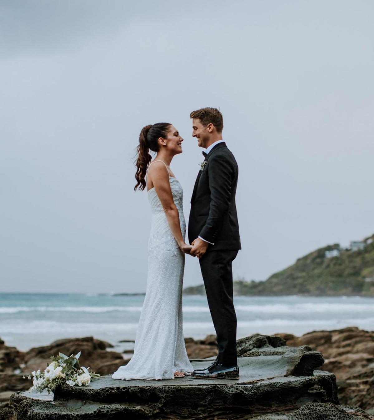 Real bride Ruby in her Darcy gown by Karen Willis Holmes. stands with her husband in front of an epic ocean background.