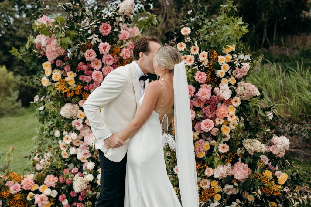 Newly wed couple, Riley and Wes, kiss in front of floral arbour at their nuptiuals. KWH real bride wears the Sage gown.
