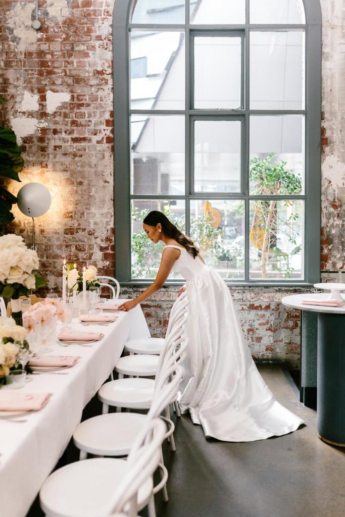 KWH real bride Anna fixing the table settings while dressed up in her Taryn Camille gown featuring U-neckline, pockets, and aline skirt.
