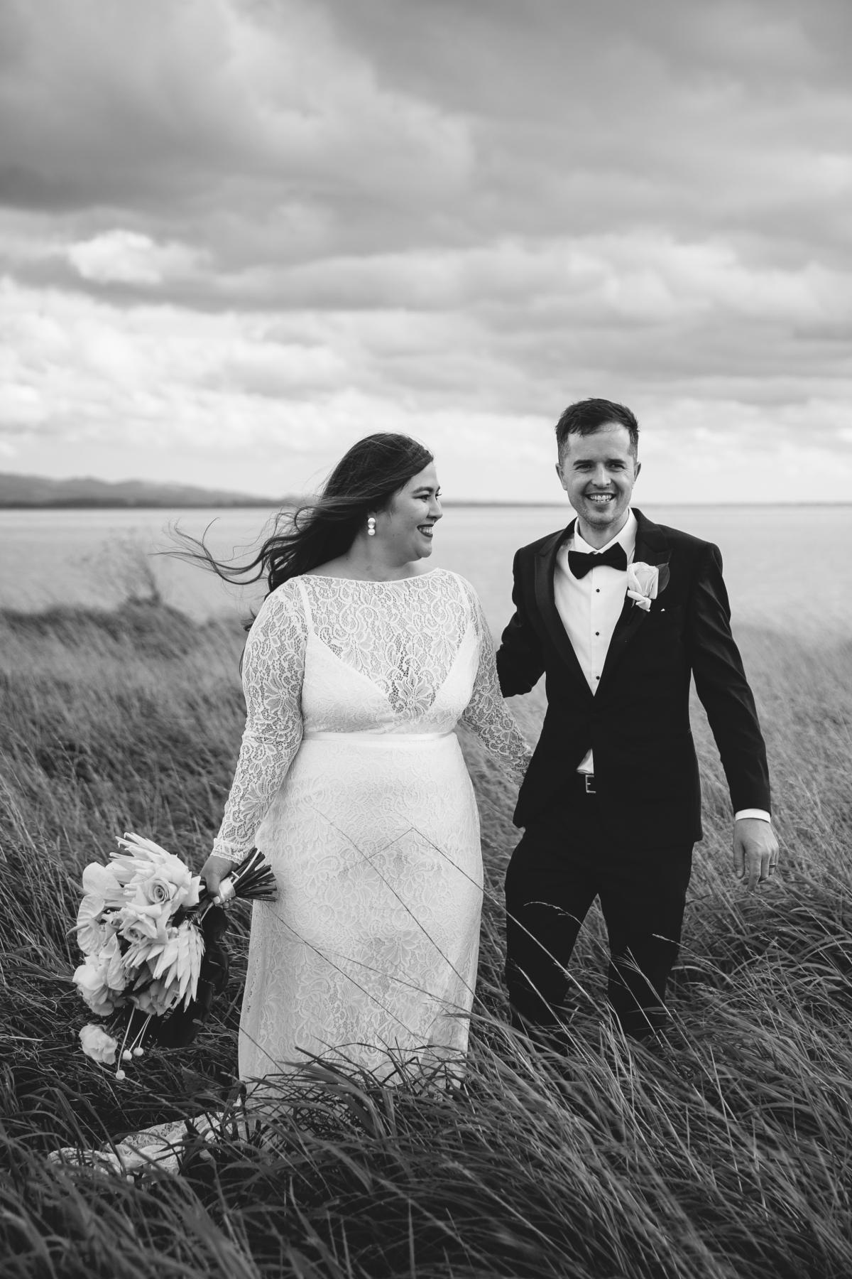 Real bride Jarna and husband Angus hold hands in dramatic Wellington landscape while she wears a long sleeve lace dress which is part of the Wild Hearts Collection.