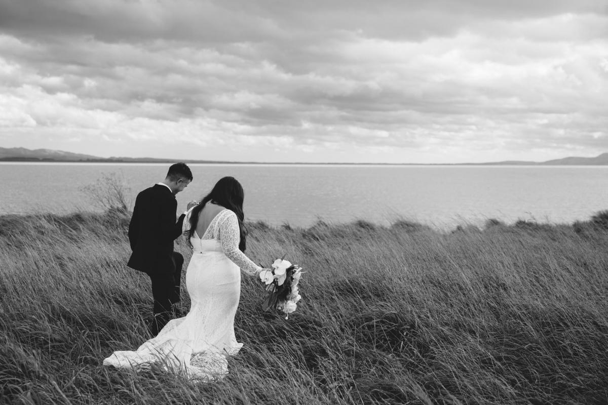 KWH bride Jarna and husband Aden walk in grass towards the sea in dramatic black and white photography showing off the stunning Karina dress with deep v back and long train.