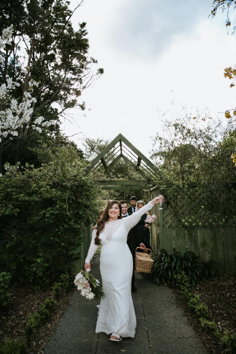 Jarna celebrates while walking out of modern outdoor wedding ceremony while looking incredible in her fitted Karina wedding dress; featuring long lace sleeves and modern lace.