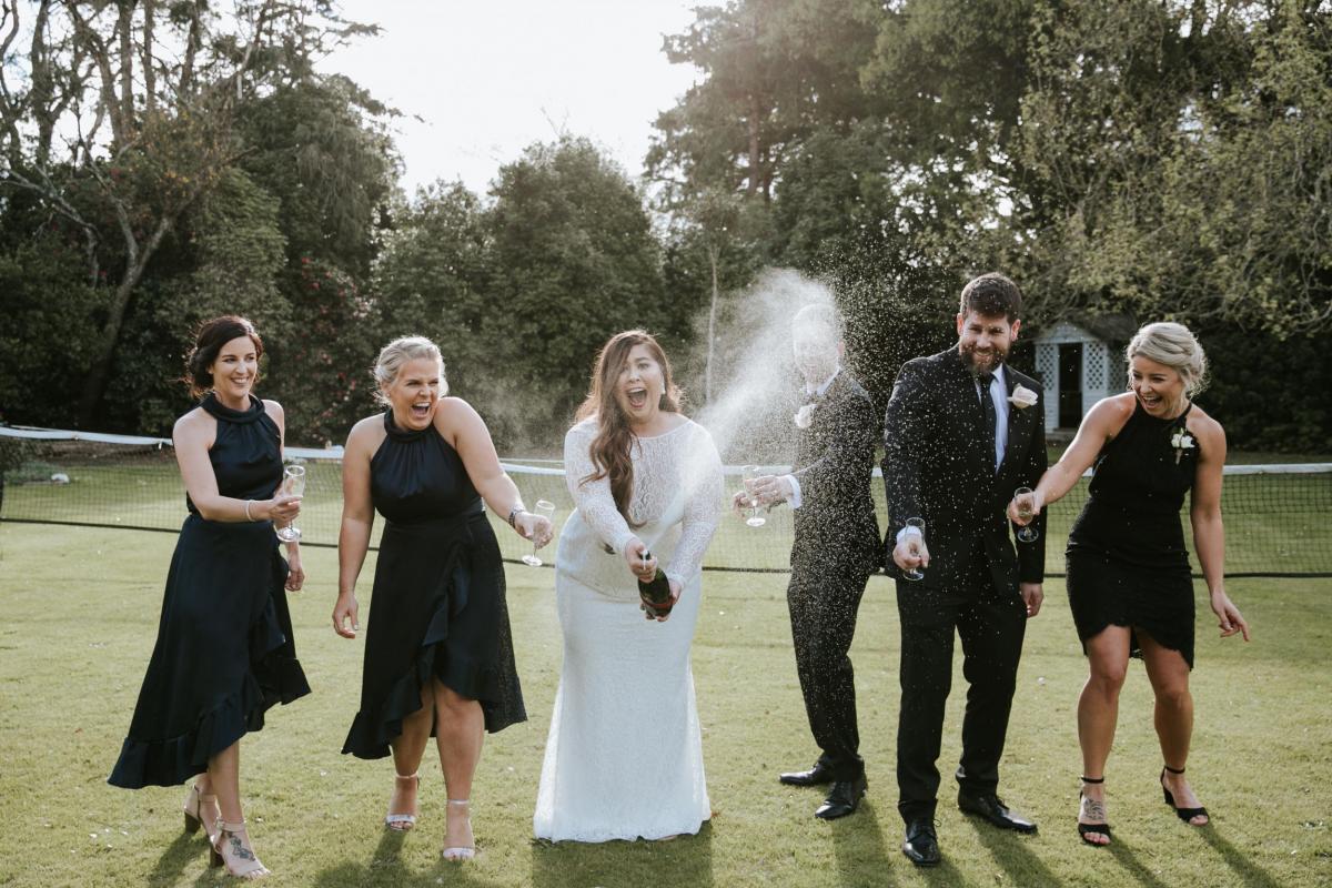 Champagne erupts from bottle as real bride Jarna, husband Aden, and wedding party celebrate the new couple in an outdoor modern cocktail hour.