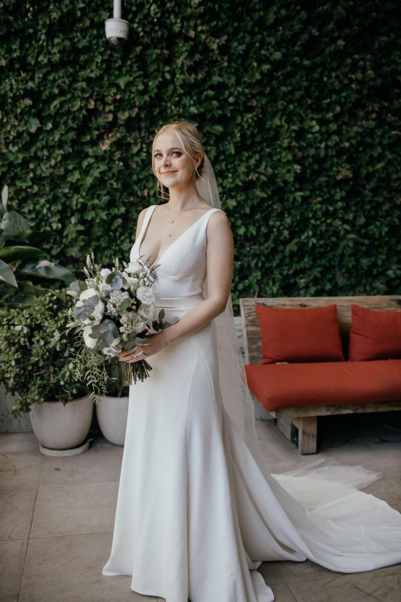 KWH Real bride Eliza stands with her white bridal bouquet in front of greenery. She wears the ivory Imogen dress by KWH.