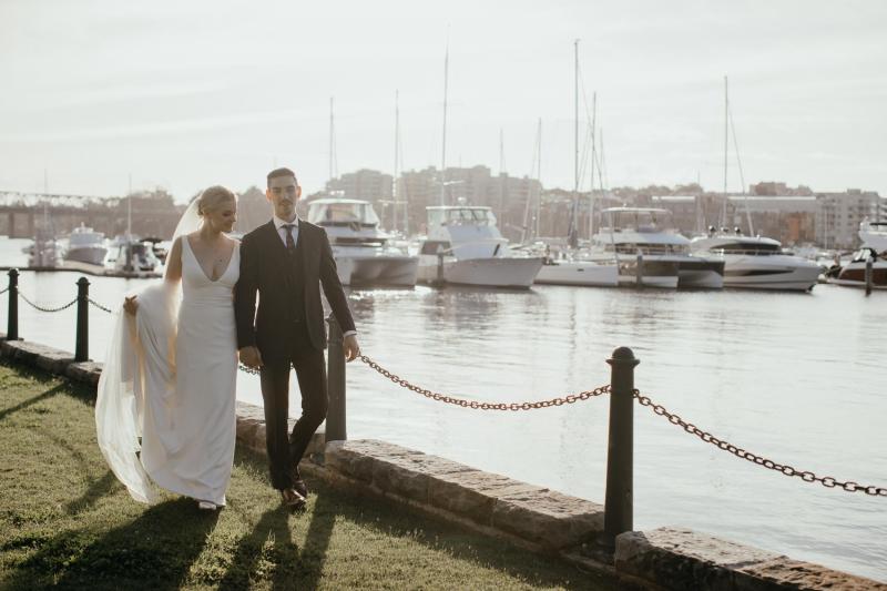 Real bride Eliza and Arien walk by the harbour with sailboats in the background. She wears the ultra sophisticated Imogen wedding gown by KWH.
