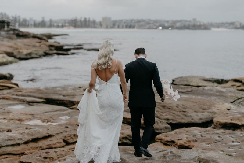 KWH real bride Nicole and Chris walking on the rocky Manly shoreline as she holds the train of her trendy Justine gown.