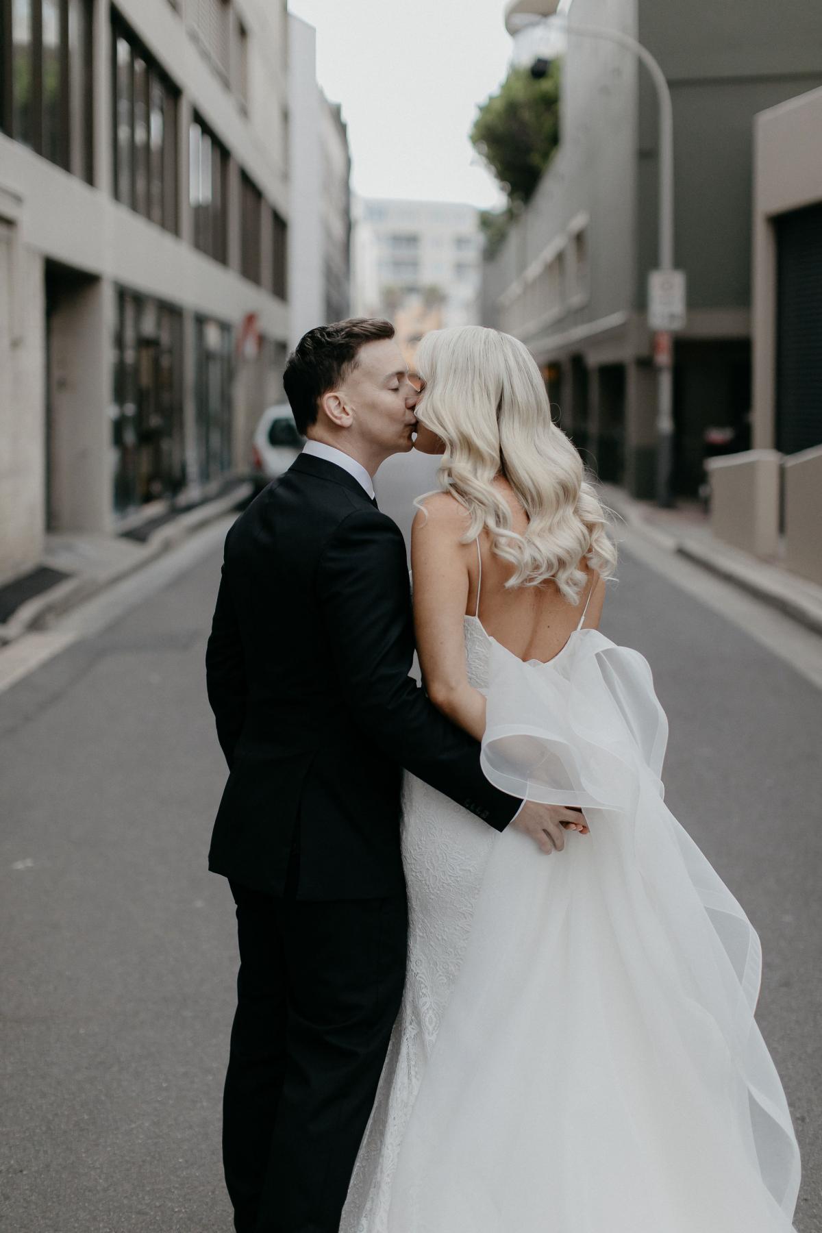 KWH real bride Nicole and Chris kiss in the middle of the street. She wears the Odette train on top of her lace Justine gown.