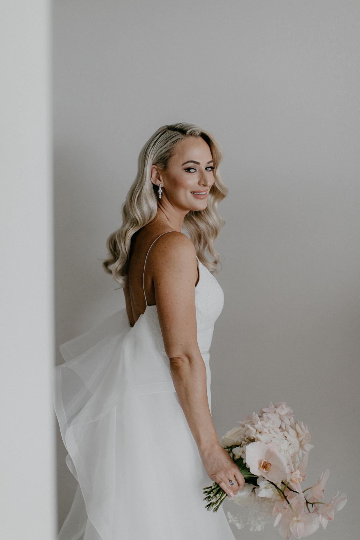 KWH real bride Nicole stands in against a gray wall for her bridal protraits wearing the trendy Justine gown with detachable Odette train and blush florals.