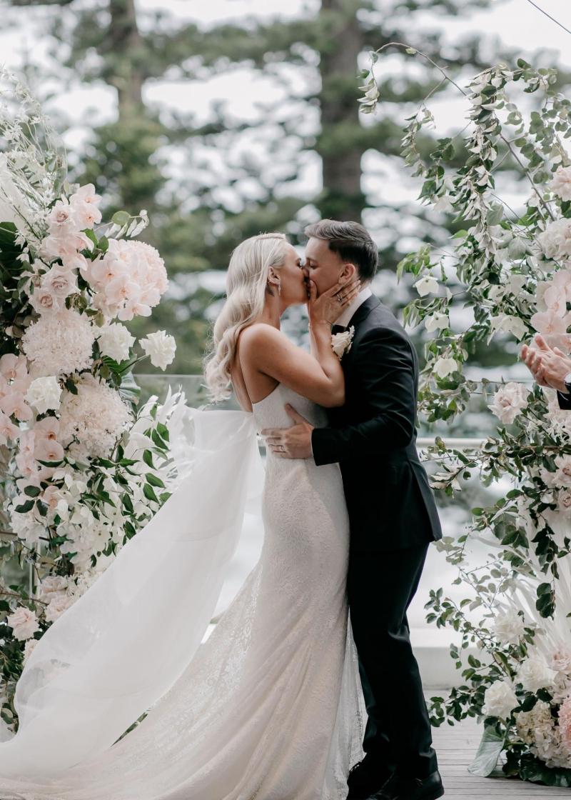 Real bride Nicole kisses her new husband Chris at the floral alter. She wears the modern lace Justine gown with Odette train from KWH.