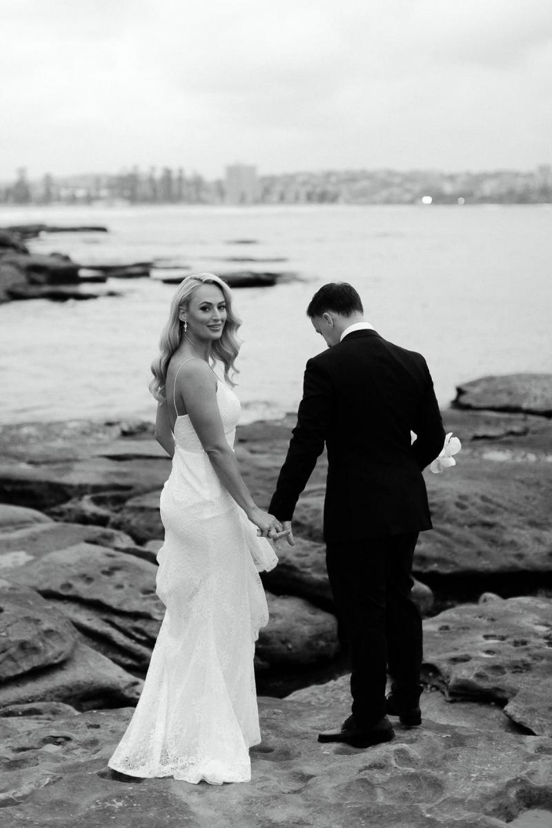 Black and white photo of real bride Nicole holding hands with her new husband on the rocky shoreline. She wears the effortless Justine gown by KWH.