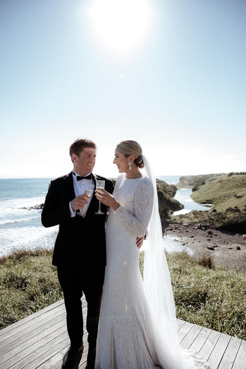 KWH real bride Hannah and Angus exchange looks while sharing a glass of champagne on the coast line. She wears the fit and flare beaded Margaretta wedding dress.