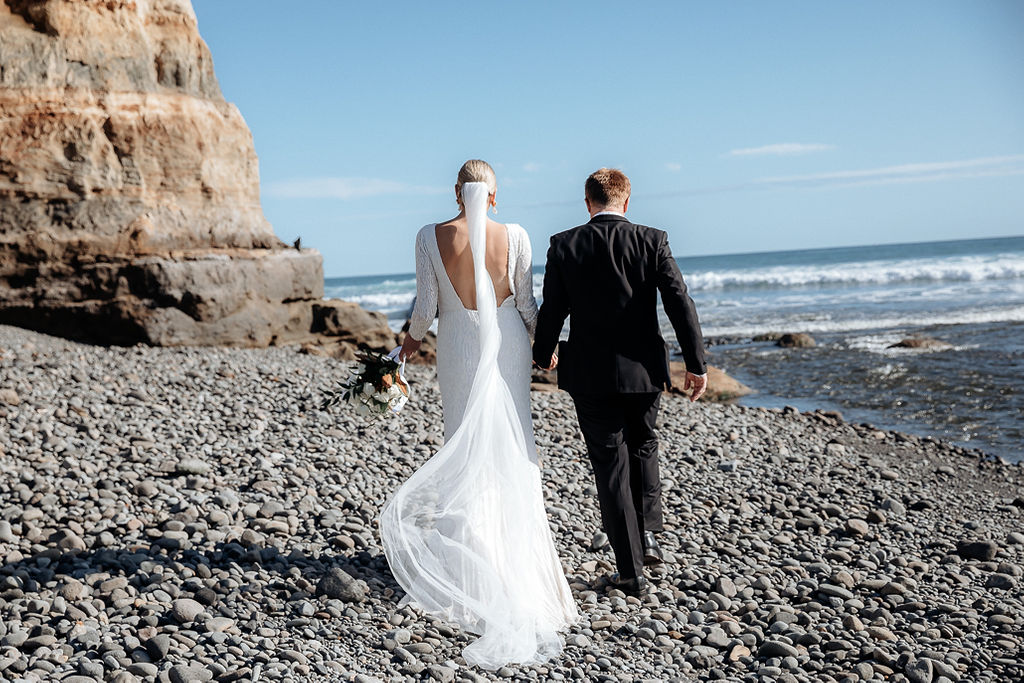 Real bride Hannah walking away with her new husband, Angus, down the shore. Her veil and Margarta dress by KWH trail behind.