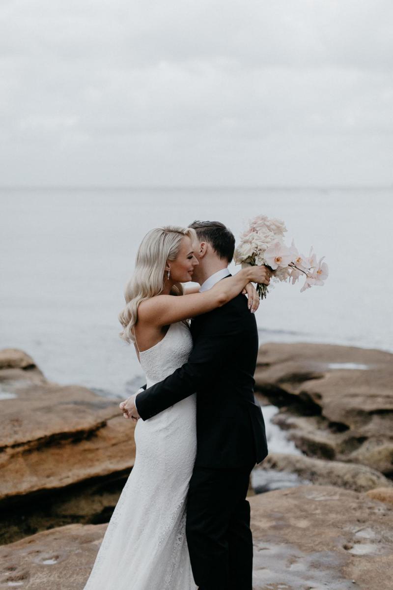 Black and white photo of real bride Nicole holding hands with her new husband on the rocky shoreline. She wears the effortless Justine gown by KWH.