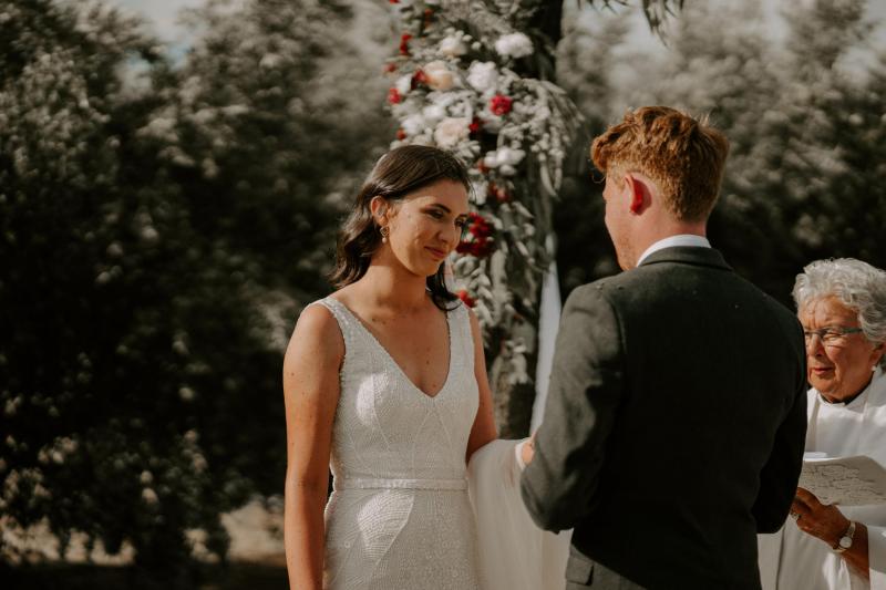 Real bride Amber and Stewart exchanging vows in from of floral arbour. She wears the beaded Olympia gown which features a v-neckline and organic inspired sequins.