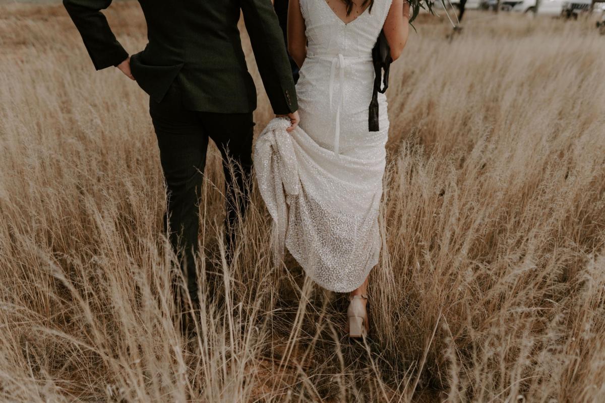 KWH real bride Amber has her beaded train of her Olympia gown carried by her new husband Stewart while walking through a field.