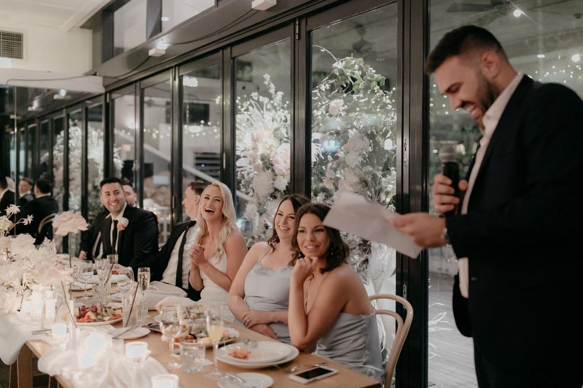 KWH real bride Nicole and Chris sit at their wedding table as the best man gives a speech. The bride wears the effortless Justine gown.